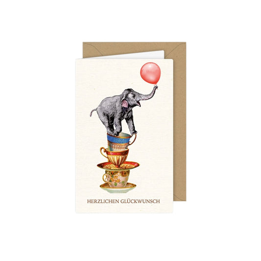 Klappkarte mit Vintage-Illustration: Ein kleiner Elefant balanciert geschickt auf einem Stapel aus gestapelten, verzierten Porzellantassen und hält einen rosa Luftballon mit seinem Rüssel. Unten steht der Schriftzug „HERZLICHEN GLÜCKWUNSCH“.