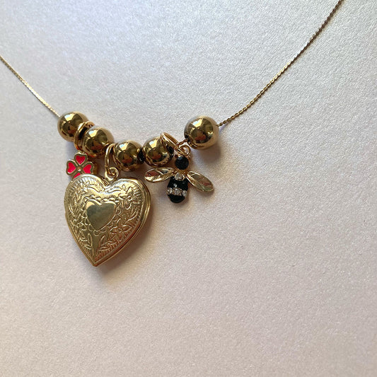 Detailaufnahme einer goldenen Halskette mit mehreren Anhängern: Ein großes, graviertes Herz-Medaillon, ein kleiner rosa Glücksklee und ein schwarz-goldener Bienen-Charm mit Flügeln, aufgereiht zwischen glänzenden Goldperlen.