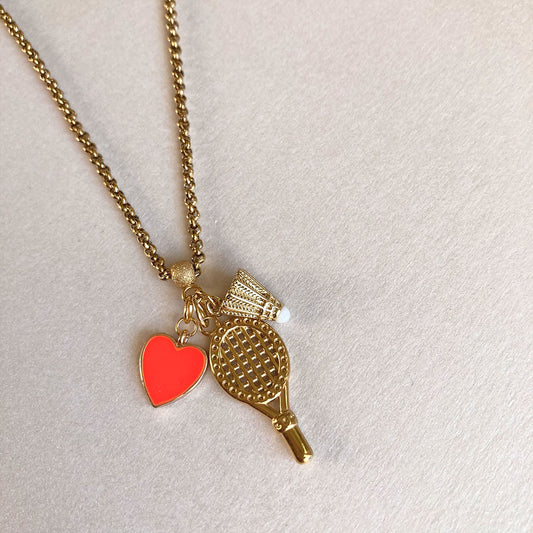 Goldene Halskette aus Edelstahl mit drei filigranen Anhängern: einem detaillierten Badminton-Schläger, einem kleinen Federball mit weißer Spitze und einem leuchtend orangefarbenen Herz-Charm, fotografiert auf einem hellen Hintergrund.