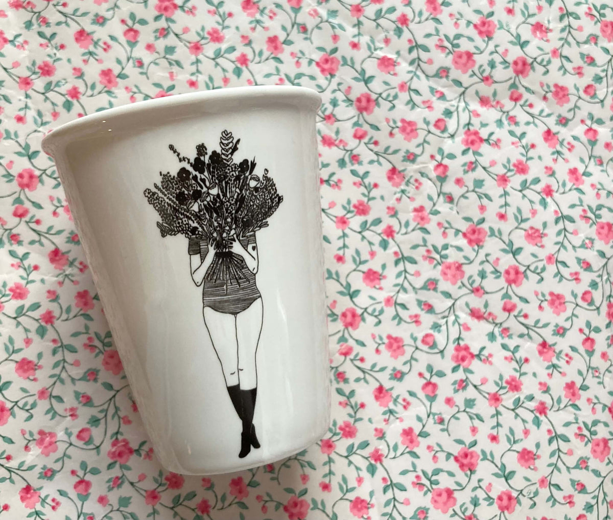 Weißer Porzellanbecher mit schwarzer Illustration einer Frau, die einen großen Blumenstrauß vor ihr Gesicht hält, vor rosafarbenem Blümchenmuster.