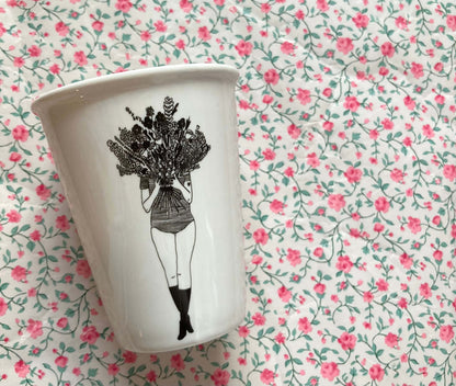 Weißer Porzellanbecher mit schwarzer Illustration einer Frau, die einen großen Blumenstrauß vor ihr Gesicht hält, vor rosafarbenem Blümchenmuster.