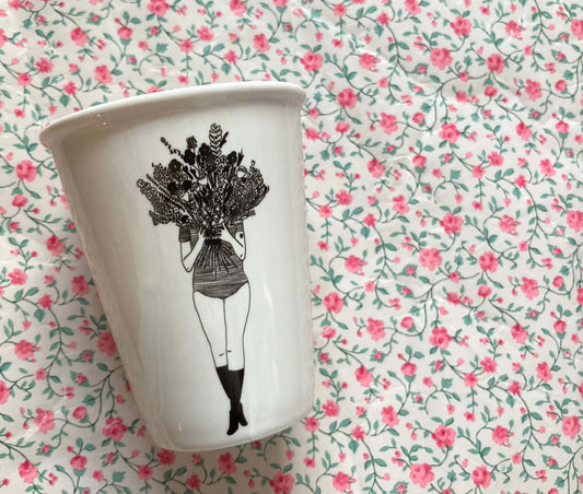 Weißer Porzellanbecher mit schwarzer Illustration einer Frau, die einen großen Blumenstrauß vor ihr Gesicht hält, vor rosafarbenem Blümchenmuster.