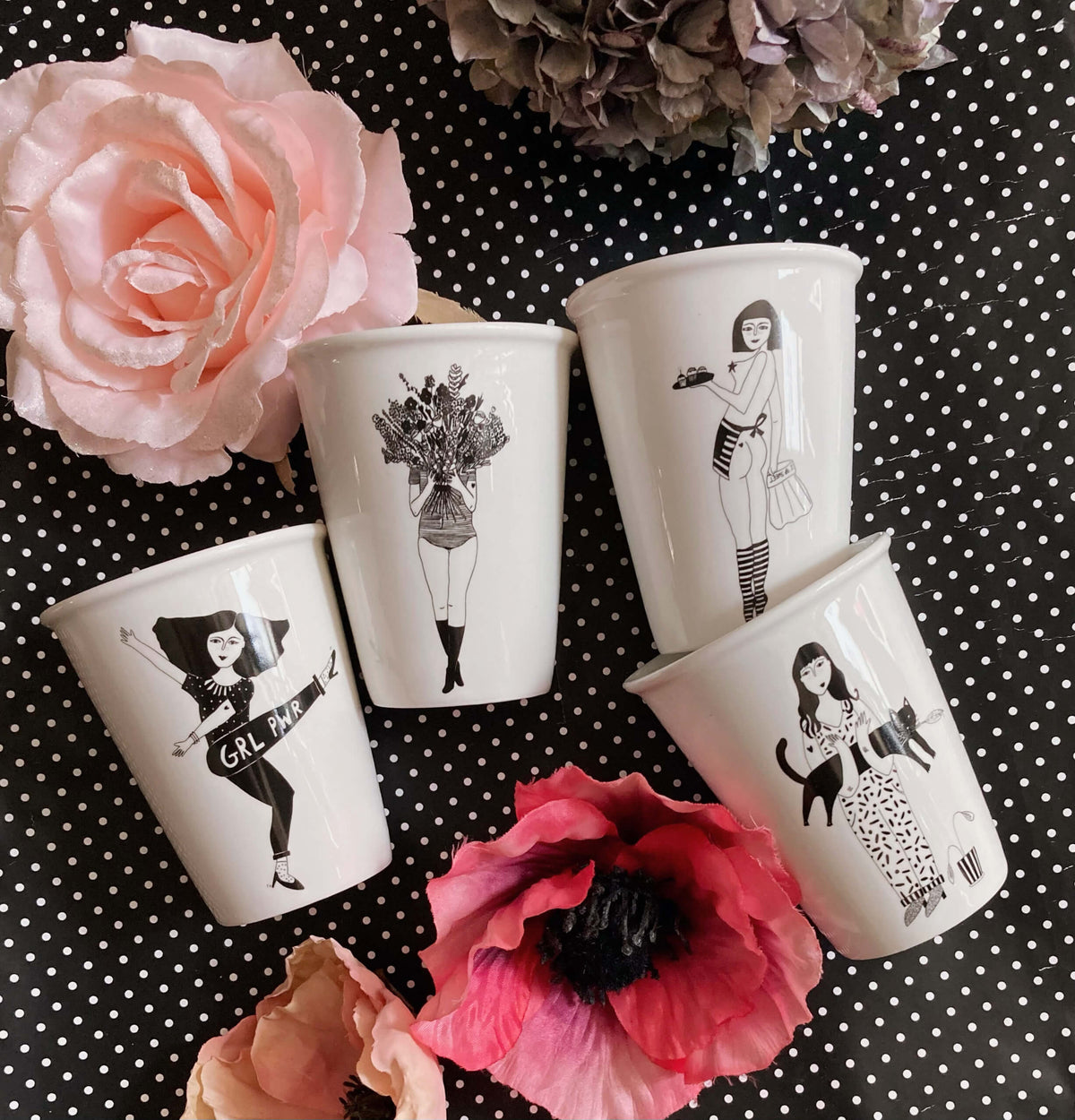 Vier weiße Porzellanbecher von helen b mit schwarzen Illustrationen – darunter Motive wie eine Tänzerin mit „GRL PWR“-Schärfe, eine Frau mit Blumenstrauß, eine Kellnerin und eine Frau mit Katze –, arrangiert auf einem schwarz-weiß gepunkteten Untergrund mit rosa Blüten.