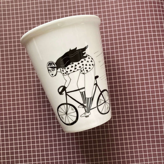 Weißer Porzellanbecher mit schwarzer Illustration einer Radfahrerin auf einem Rennrad vor dunklem Karomuster.