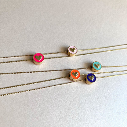 Fünf goldene Halsketten sind kreuz und quer auf einem hellen, glatten Hintergrund ausgelegt. Die runden Anhänger mit bunten Emaille-Details und goldenen Herz-Motiven stehen im Fokus. Man sieht deutlich die seitliche Öse der Anhänger, durch die die Kette verläuft. Die Farben wirken lebendig und sommerlich.