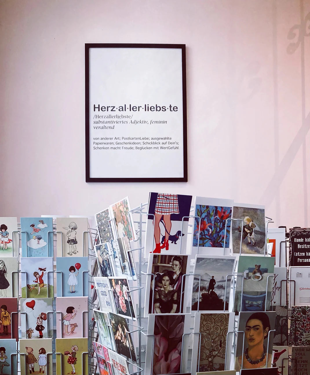 Innenansicht des Ladengeschäfts „Herzallerliebste“ mit gut gefüllten Postkartenständern voller künstlerischer Motive und Illustrationen. An der Wand hängt ein markantes Poster, das den Ladennamen „Herz·al·ler·liebs·te“ wie einen Lexikoneintrag erklärt und Werte wie „PostkartenLiebe“, „ausgewählte Papierwaren“ und „Geschenkideen“ hervorhebt. Die Szene vermittelt eine einladende, nostalgische Atmosphäre für Liebhaber von hochwertiger Papeterie und besonderen Geschenken.
