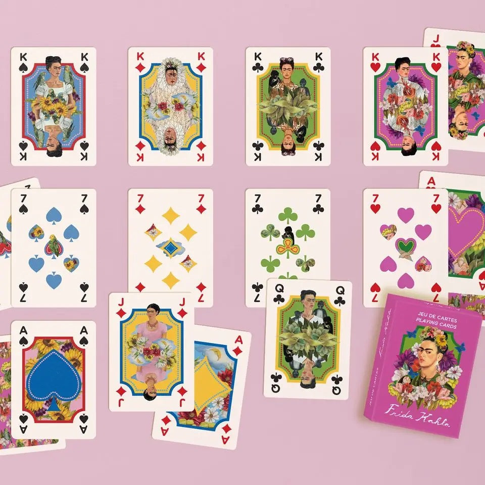 Mehrere Spielkarten des Frida Kahlo Decks sind übersichtlich in Reihen auf einem hellrosa Hintergrund ausgelegt. Die obere Reihe zeigt die vier Könige, dargestellt als Frida Kahlo in verschiedenen traditionellen Kleidern und Farbschemata (Pik, Karo, Kreuz, Herz). Die mittleren Reihen zeigen kunstvoll gestaltete Zahlenkarten (Siebenen) und Buben. Unten rechts liegt die magentafarbene Verpackung neben einer Dame-Karte.