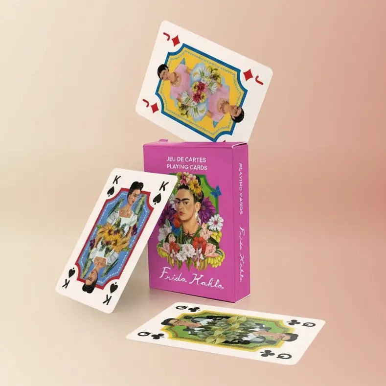 Ein stilvolles Produktfoto vor einem sanften, beigefarbenen Farbverlauf. Die magentafarbene Frida Kahlo Spielkartenbox steht aufrecht. Eine Bube-Karte balanciert magisch auf der Oberkante der Box, während eine König-Karte schräg daran lehnt. Eine weitere Karte liegt flach im Vordergrund auf dem Boden, was dem Arrangement eine elegante Tiefe verleiht.