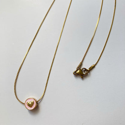 Eine sehr feine, goldene Gliederkette flach liegend auf hellen Untergrund. Der runde Anhänger hat eine zartrosa Emaille-Füllung, in deren Mitte ein kleines, plastisches, goldenes Herz eingearbeitet ist.