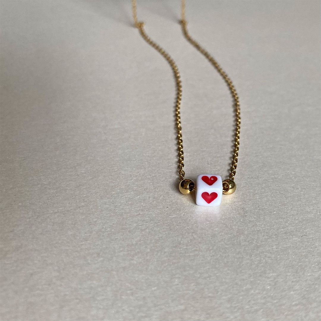 Zarte goldene Gliederkette mit einem kleinen weißen Würfel in der Mitte, auf dem ein rotes Herz abgebildet ist.