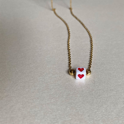 Zarte goldene Gliederkette mit einem kleinen weißen Würfel in der Mitte, auf dem ein rotes Herz abgebildet ist.