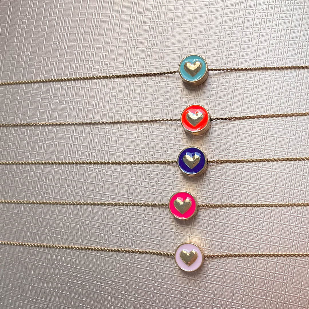 Eine Nahaufnahme von fünf feingliedrigen, goldfarbenen Ketten, die parallel untereinander liegen. Jede Kette hat einen runden, münzähnlichen Anhänger mit einem farbigen Emaille-Inlay und einem erhabenen goldenen Herz in der Mitte. Die Farben der Anhänger von oben nach unten sind: Hellblau, Orange-Rot, Dunkelblau, Neon-Pink und Pastellrosa.