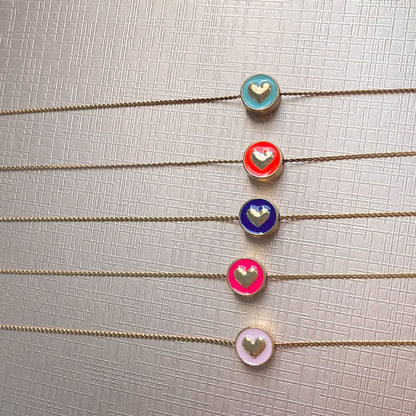 Eine Nahaufnahme von fünf feingliedrigen, goldfarbenen Ketten, die parallel untereinander liegen. Jede Kette hat einen runden, münzähnlichen Anhänger mit einem farbigen Emaille-Inlay und einem erhabenen goldenen Herz in der Mitte. Die Farben der Anhänger von oben nach unten sind: Hellblau, Orange-Rot, Dunkelblau, Neon-Pink und Pastellrosa.