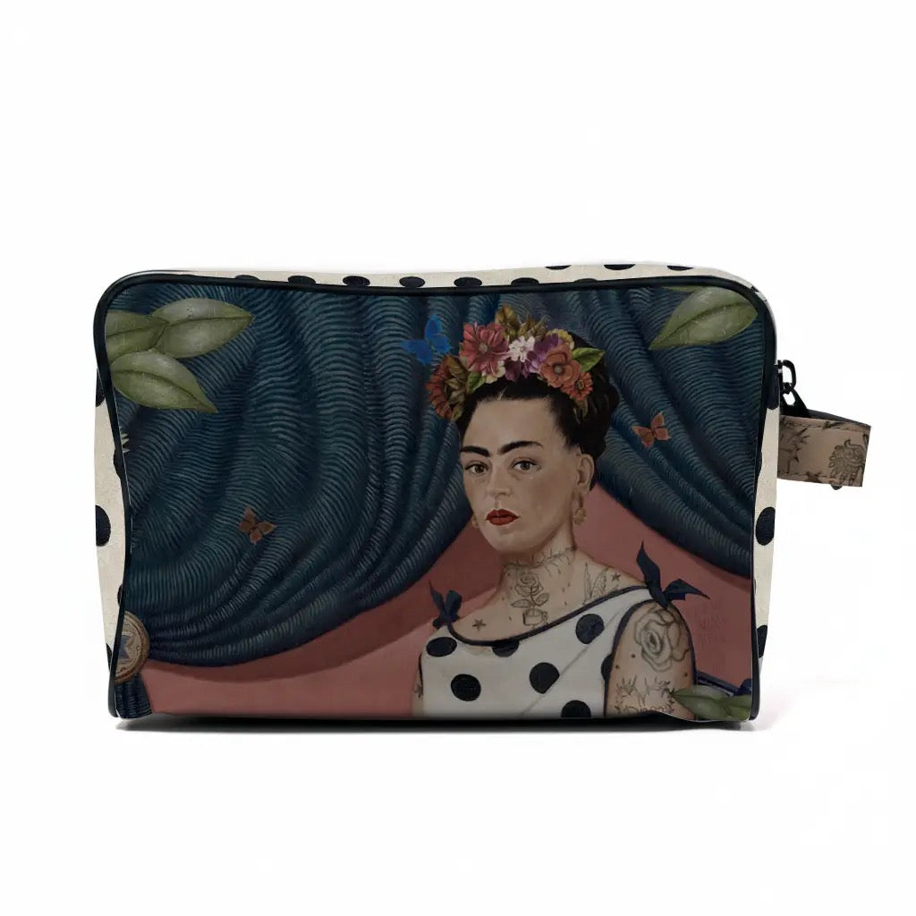 Vorderseite der voglio bene Kulturtasche mit einem kunstvollen Porträt von Frida Kahlo. Sie trägt einen bunten Blumenkranz im Haar, ein gepunktetes Kleid und feine Tattoos an Hals und Schultern. Der Hintergrund zeigt einen dunkelblauen, drapierten Vorhang mit Schmetterlingen.