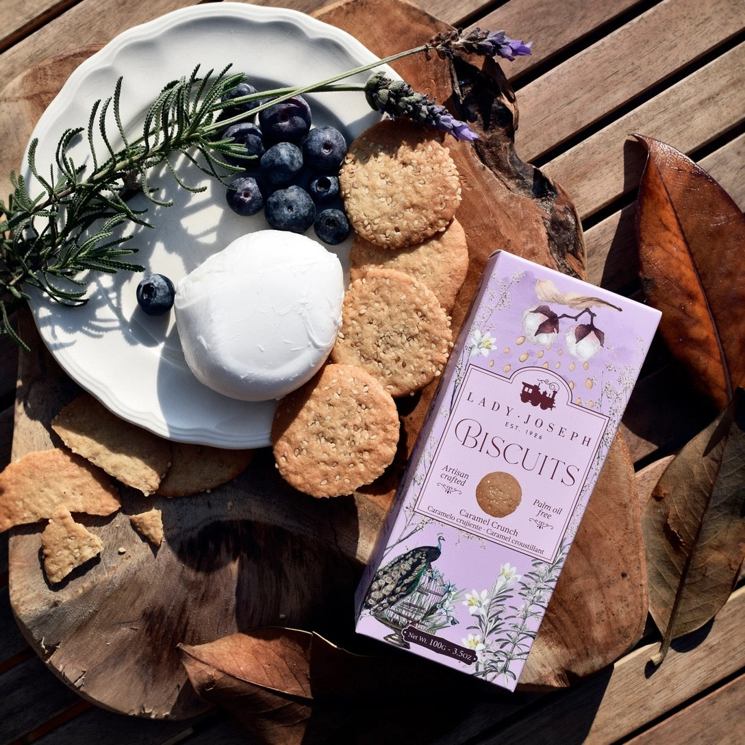 Auf einem weißen Teller liegen Frischkäse, Heidelbeeren, Haferkekse und ein Zweig Rosmarin, daneben steht eine lilafarbene Packung Lady Joseph Biscuits auf einem Holzbrett mit getrockneten Blättern.
