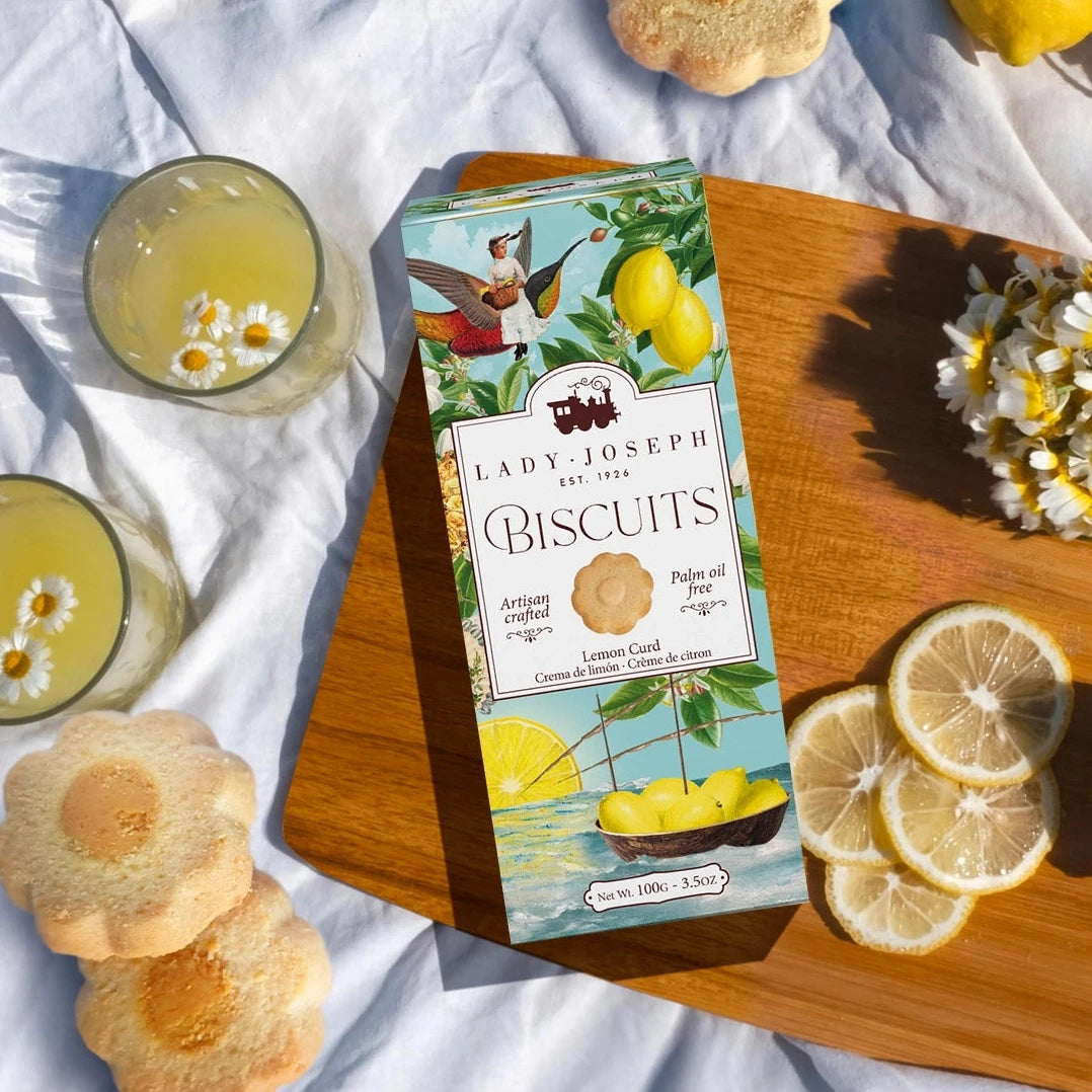 Holzbrett mit geschnittenen Zitronenscheiben, einer Packung Lady Joseph Lemon Curd Biscuits, zwei Gläsern mit Getränk und Kamillenblüten, mehreren Blütenförmigen Keksen, einem Blumenstrauß aus Kamillen und zwei ganzen Zitronen auf weißem Tuch.