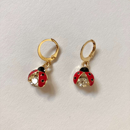 Goldene Creolen-Ohrringe mit Marienkäfer-Anhängern. Die Marienkäfer haben rote Flügel mit schwarzen Punkten und einen großen, funkelnden Kristall-Körper. 
