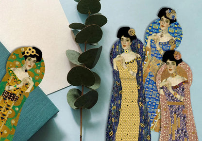 Flachbild-Arrangement von vier figürlich gestanzten Gustav-Klimt-Lesezeichen auf einem hellblauen Hintergrund. Neben den Lesezeichen liegt ein grüner Eukalyptuszweig, ein weißes Papierblatt und ein petrolfarbenes Buch. Die Lesezeichen zeigen das Porträt einer eleganten Frau im Jugendstil, gekleidet in gemusterten Roben in Grün, Gelb-Blau, leuchtendem Blau und Altrosa.