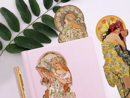 Ein zartrosa Notizbuch auf hellem Hintergrund, flankiert von einem grünen Zweig und einem goldenen Stift. Ein Lesezeichen einer seitlich blickenden Renaissance-Frau im dunkel gemusterten Kleid mit weißen Rosen in den Händen liegt auf dem Buch, während im Hintergrund zwei weitere Botticelli-Figuren zu sehen sind.