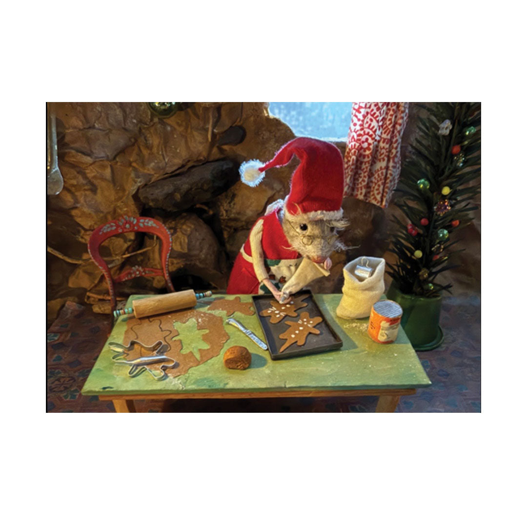 Eine gefilzte Maus in Weihnachtskleidung mit roter Mütze und Brille backt Plätzchen an einem grünen Holztisch. Auf dem Tisch liegen ausgerollter Teig, ein Nudelholz, Plätzchenausstecher und ein Backblech mit mauszackigen Lebkuchenmännern, die gerade mit weißem Guss dekoriert werden. Im Hintergrund befinden sich eine Steinwand und ein kleiner Weihnachtsbaum.