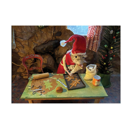 Eine gefilzte Maus in Weihnachtskleidung mit roter Mütze und Brille backt Plätzchen an einem grünen Holztisch. Auf dem Tisch liegen ausgerollter Teig, ein Nudelholz, Plätzchenausstecher und ein Backblech mit mauszackigen Lebkuchenmännern, die gerade mit weißem Guss dekoriert werden. Im Hintergrund befinden sich eine Steinwand und ein kleiner Weihnachtsbaum.