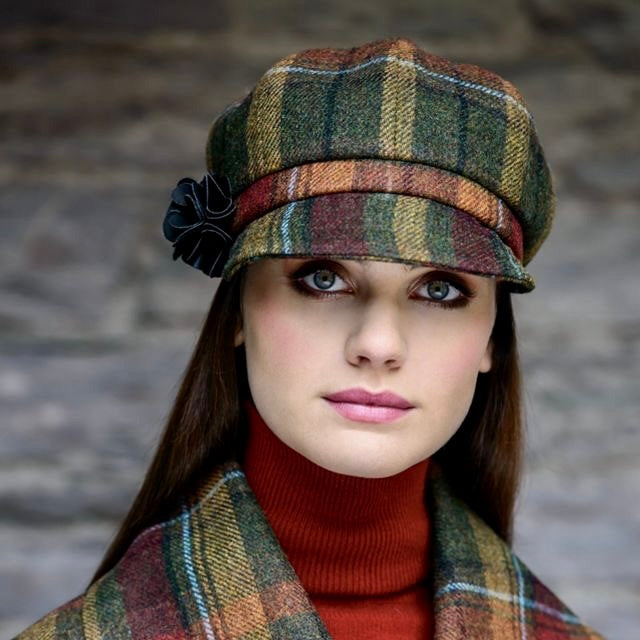 Frau trägt karierten Mantel und passende Schirmmütze in Grün-, Rot- und Beigetönen mit schwarzer Blume als Accessoire am Hut.