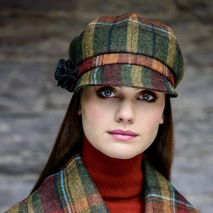 Frau trägt karierten Mantel und passende Schirmmütze in Grün-, Rot- und Beigetönen mit schwarzer Blume als Accessoire am Hut.