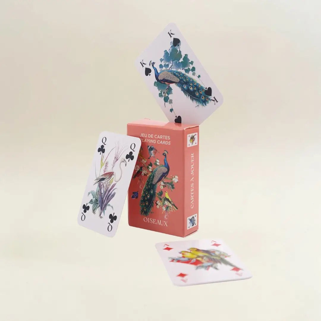Eine kreative Produktpräsentation vor einem neutralen, hellen Hintergrund. Auf der lachsfarbenen „Oiseaux“-Kartenbox balanciert an der oberen Kante eine König-Karte mit einem Pfauenmotiv. Links lehnt eine Kreuz-Dame, die einen Flamingo im Schilf zeigt, schräg an der Box. Eine weitere Karte mit zwei gelb-grünen Sittichen liegt flach im Vordergrund.