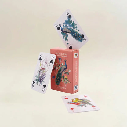 Eine kreative Produktpräsentation vor einem neutralen, hellen Hintergrund. Auf der lachsfarbenen „Oiseaux“-Kartenbox balanciert an der oberen Kante eine König-Karte mit einem Pfauenmotiv. Links lehnt eine Kreuz-Dame, die einen Flamingo im Schilf zeigt, schräg an der Box. Eine weitere Karte mit zwei gelb-grünen Sittichen liegt flach im Vordergrund.