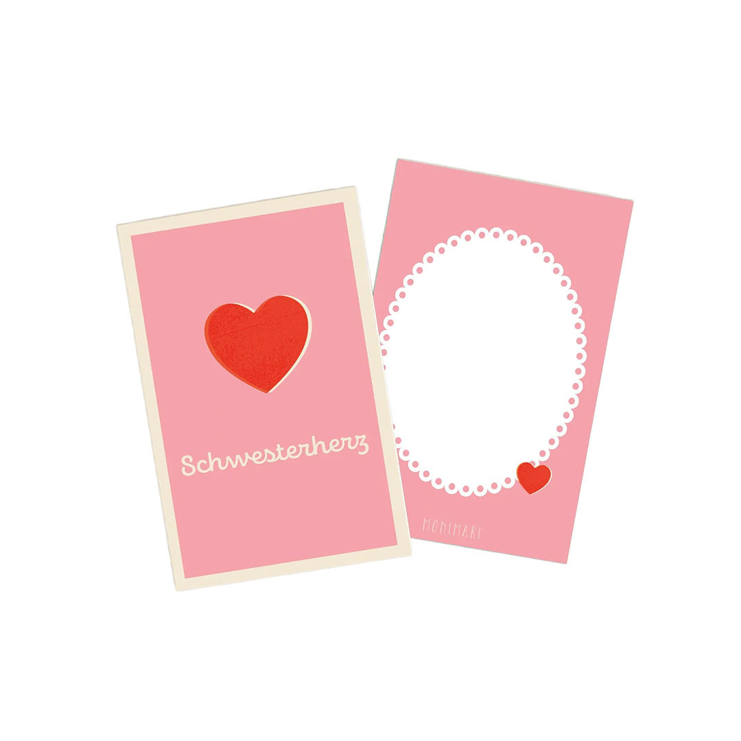 Minikarte von Monimari in Rosa. Vorderseite: Ein rotes Herz im Zentrum und der geschwungene Schriftzug „Schwesterherz“ darunter. Rückseite: Rosafarbener Hintergrund mit weißem, geperltem Oval-Rahmen und einem kleinen roten Herz unten rechts.