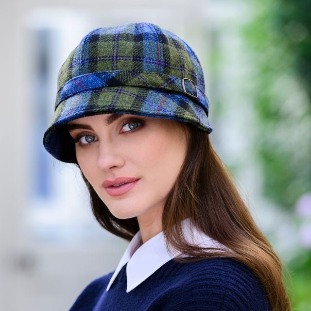 Eine Frau mit blau grünfarbenem, kariertem Flapper Hut, Dunkles Oberteil mit weißem Kragen vor grünem Laub im Hintergrund.