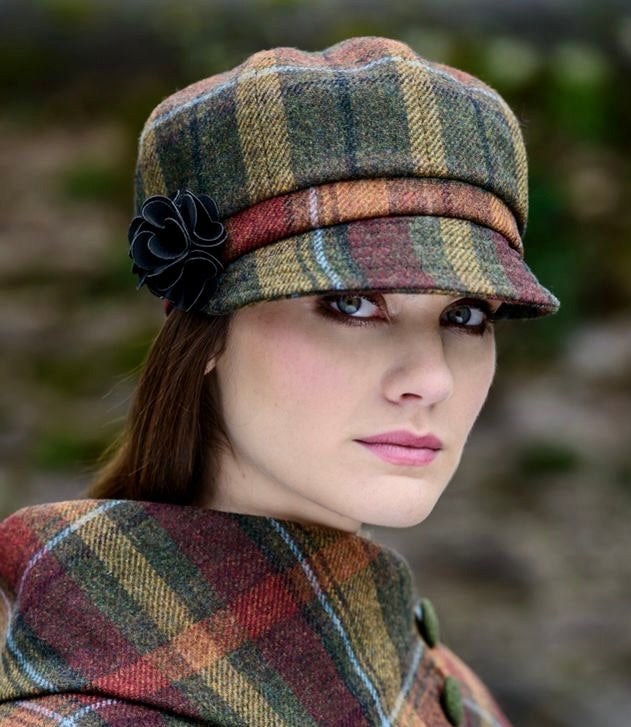 Frau  trägt karierten Mantel und passende Schirmmütze in Grün-, Rot- und Beigetönen mit schwarzer Blume als Accessoire am Hut.