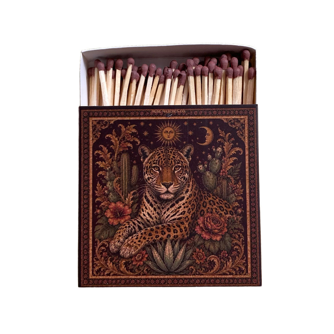 Vorderseite einer braunen Muse Matches Co. Streichholzschachtel mit einer barocken Illustration eines Leoparden zwischen Kakteen und Zündhölzern mit braunen Köpfen.
