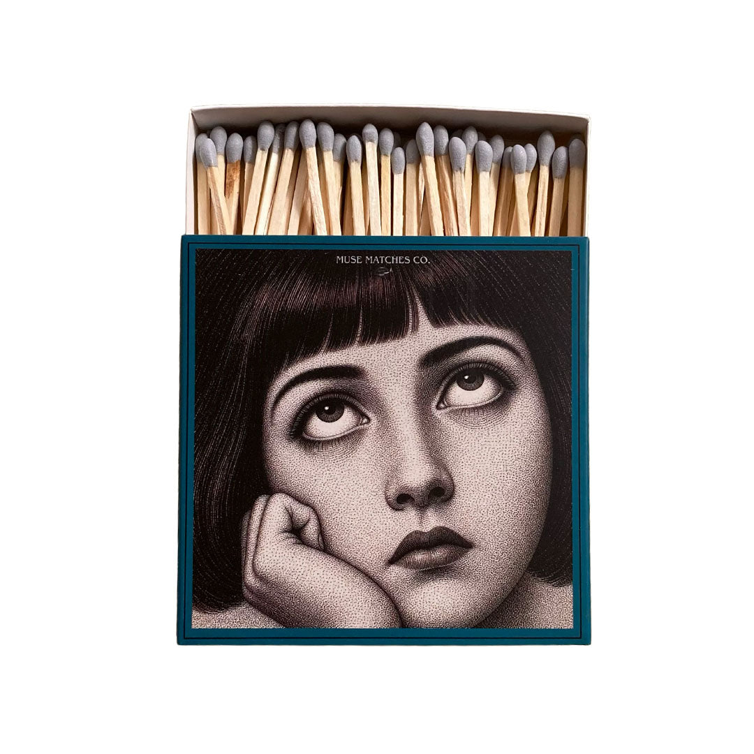 Vorderseite einer blauen Muse Matches Co. Streichholzschachtel mit dem grafischen Porträt einer nach oben blickenden Frau und grauen Streichhölzern.