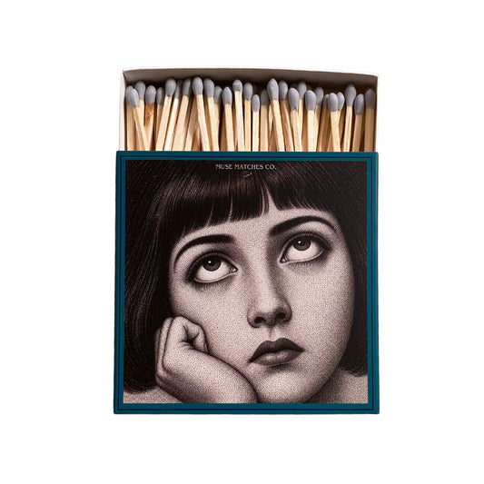 Vorderseite einer blauen Muse Matches Co. Streichholzschachtel mit dem grafischen Porträt einer nach oben blickenden Frau und grauen Streichhölzern.