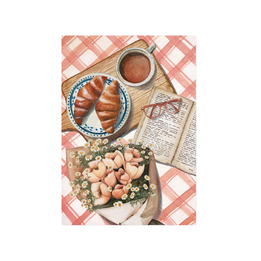 Zwei Croissants auf blau-weißem Teller, Tasse Kaffee, aufgeschlagenes Buch mit roter Brille und Blumenstrauß auf rot-weiß kariertem Tischtuch.