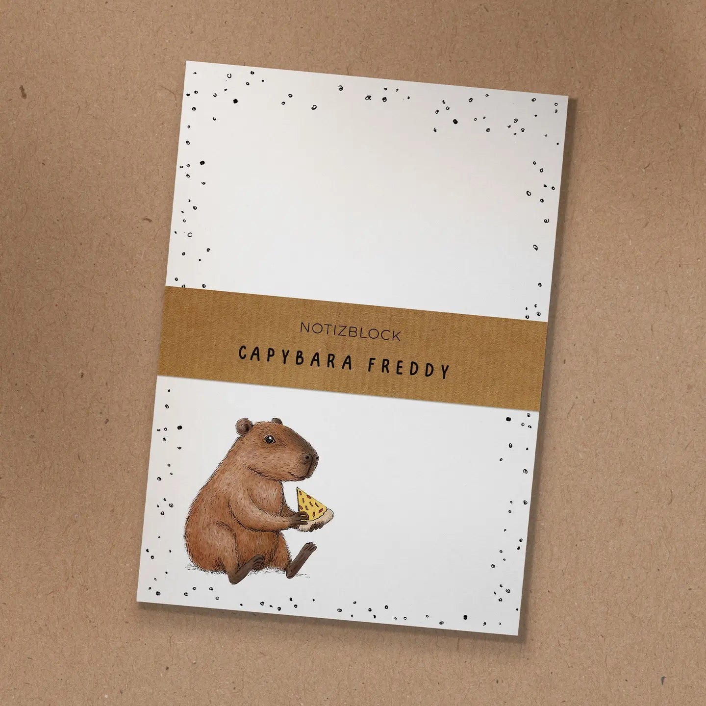 Notizblock mit Kraftpapier-Banderole und handgezeichneter Illustration von Capybara Freddy, das ein Stück Pizza isst.