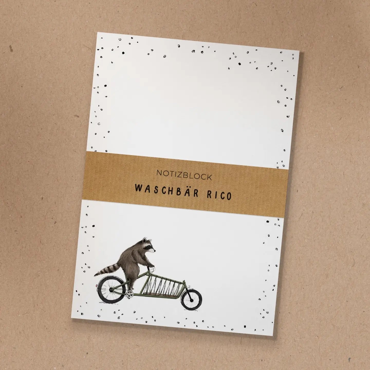 Notizblock mit Kraftpapier-Banderole und handgezeichneter Illustration von Waschbär Rico auf einem grünen Lastenrad.