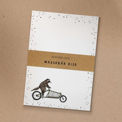 Notizblock mit Kraftpapier-Banderole und handgezeichneter Illustration von Waschbär Rico auf einem grünen Lastenrad.