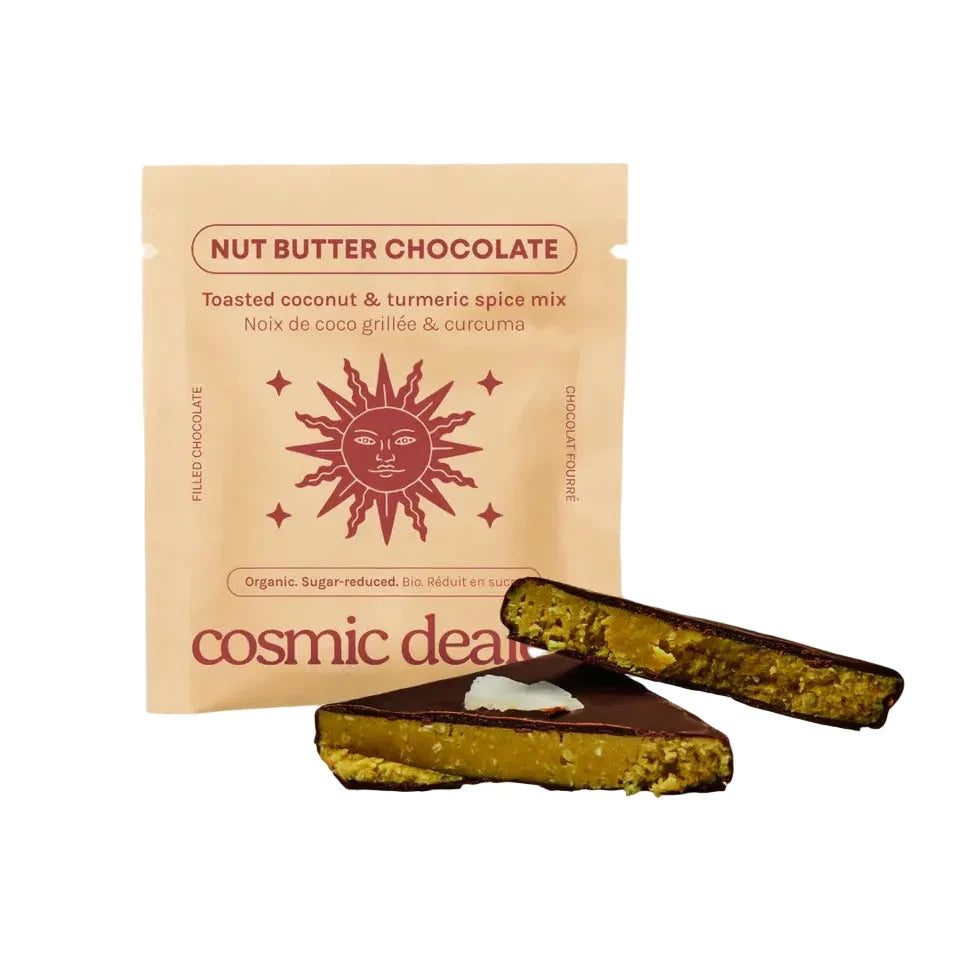 Eine hellbraune Verpackung der Sorte „Toasted coconut & turmeric spice mix“ mit einer roten Sonnen-Illustration. Davor liegen zwei angeschnittene Schokoladenstücke, die eine goldgelbe Kokos-Kurkuma-Füllung offenbaren. Der Markenname „cosmic dealer“ ist unten auf der Packung zu sehen.