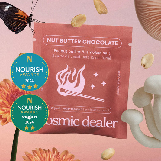 Künstlerische Inszenierung der Schokoladenverpackung in einem surrealen Setting. Das Produkt ist umgeben von schwebenden Erdnüssen, einer orangefarbenen Blume, weißen Pilzen und einem Schmetterling auf einem weichen, rosa-verlaufenden Hintergrund. Links sind die „Nourish Awards 2024“-Logos für Qualität und vegane Ernährung zu sehen.