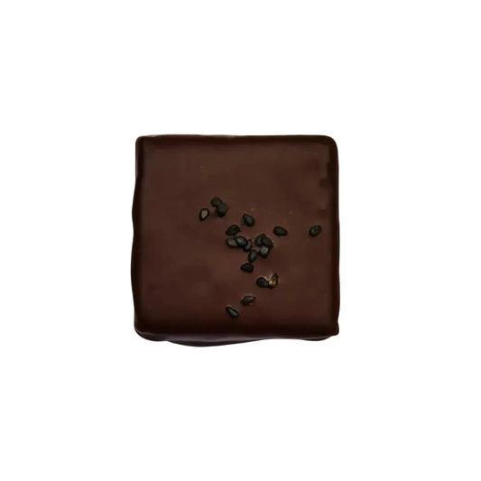 Eine quadratische Zartbitterschokolade aus der Vogelperspektive auf hellem beigem Hintergrund. In der Mitte der glatten Oberfläche sind kleine schwarze Sesamsamen dekorativ verstreut.