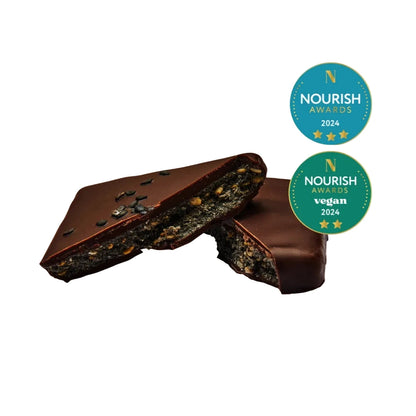 Ein in zwei Hälften gebrochenen Schokoriegel. Der Querschnitt zeigt eine reichhaltige, dunkle Füllung mit sichtbaren schwarzen Sesamsamen und Gewürzen. Im Hintergrund sind erneut die zwei „Nourish Awards 2024“-Auszeichnungen zu sehen.