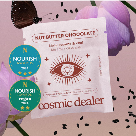Eine kunstvolle Produktinszenierung der „Black Sesame & Chai“ Nut Butter Chocolate. Die weiße Verpackung mit einem mystischen Augen-Symbol liegt vor einem Hintergrund aus lila Blüten, einem Schmetterling und verstreuten Sesamsamen. Zwei gold-blaue „Nourish Awards 2024“-Siegel sind prominent abgebildet.