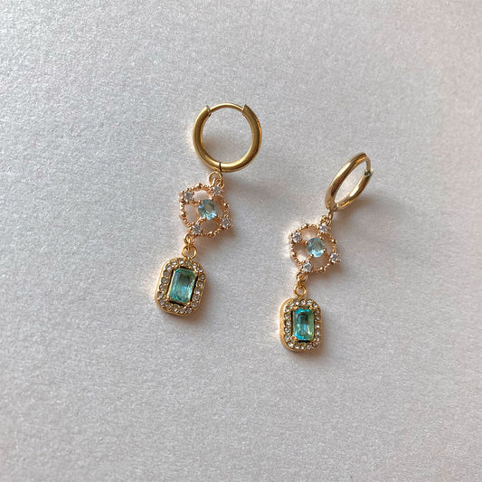 Elegante, goldene Drop-Ohrringe mit hellblauen Kristallen. Ein rundes Ornament-Element mit Zirkonia verbindet die Creole mit einem rechteckigen aquamarinen Stein im Baguette-Schliff in einer Pavé-Fassung.