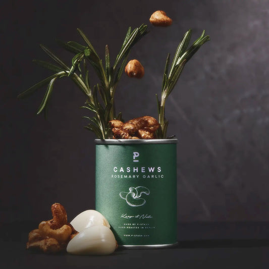 Dunkelgrüne P-Stash Dose, aus der frische Rosmarinzweige und gewürzte Cashews herausragen, flankiert von frischen Knoblauchzehen auf dunklem Untergrund.