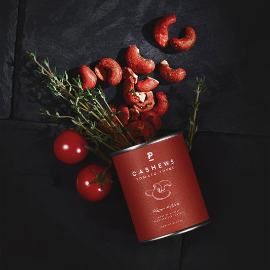 Dose mit Cashew-Nüssen, Tomate und Thymian, daneben zwei Tomaten und Thymianzweige auf dunklem Untergrund
