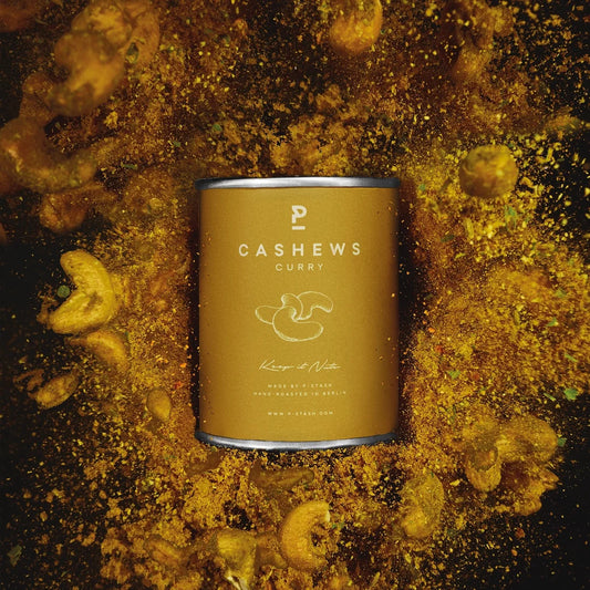 Zentrierte gelb-braune P-Stash Dose mit der Aufschrift „Cashews Curry“, umgeben von einer dynamischen Explosion aus Currypulver und fliegenden Cashewnüssen vor dunklem Hintergrund.