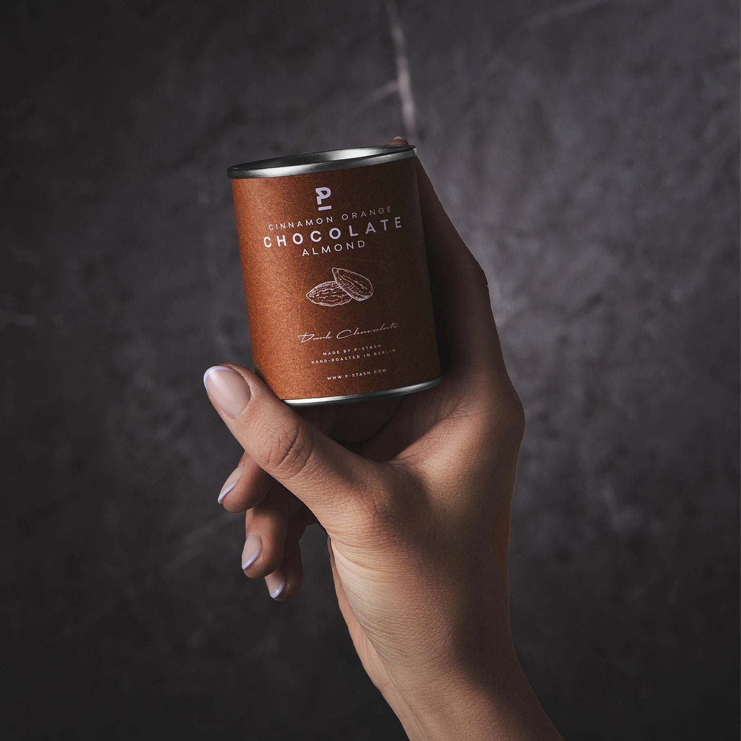 Eine Hand hält eine braune P-Stash Dose mit der Aufschrift „Cinnamon Orange Chocolate Almond“ vor einer dunkelgrau strukturierten Wand.