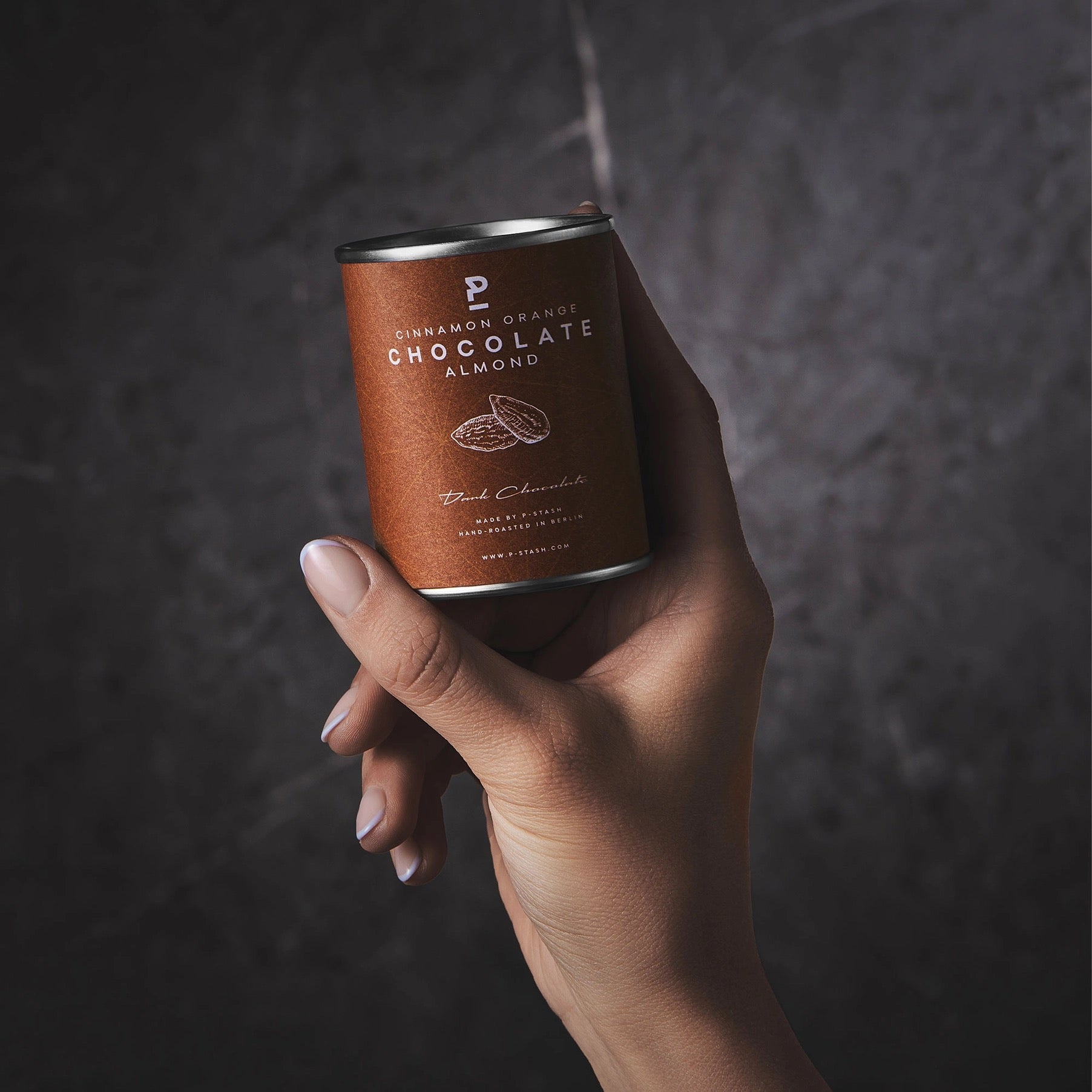 Eine Hand hält eine braune P-Stash Dose mit der Aufschrift „Cinnamon Orange Chocolate Almond“ vor einer dunkelgrau strukturierten Wand.